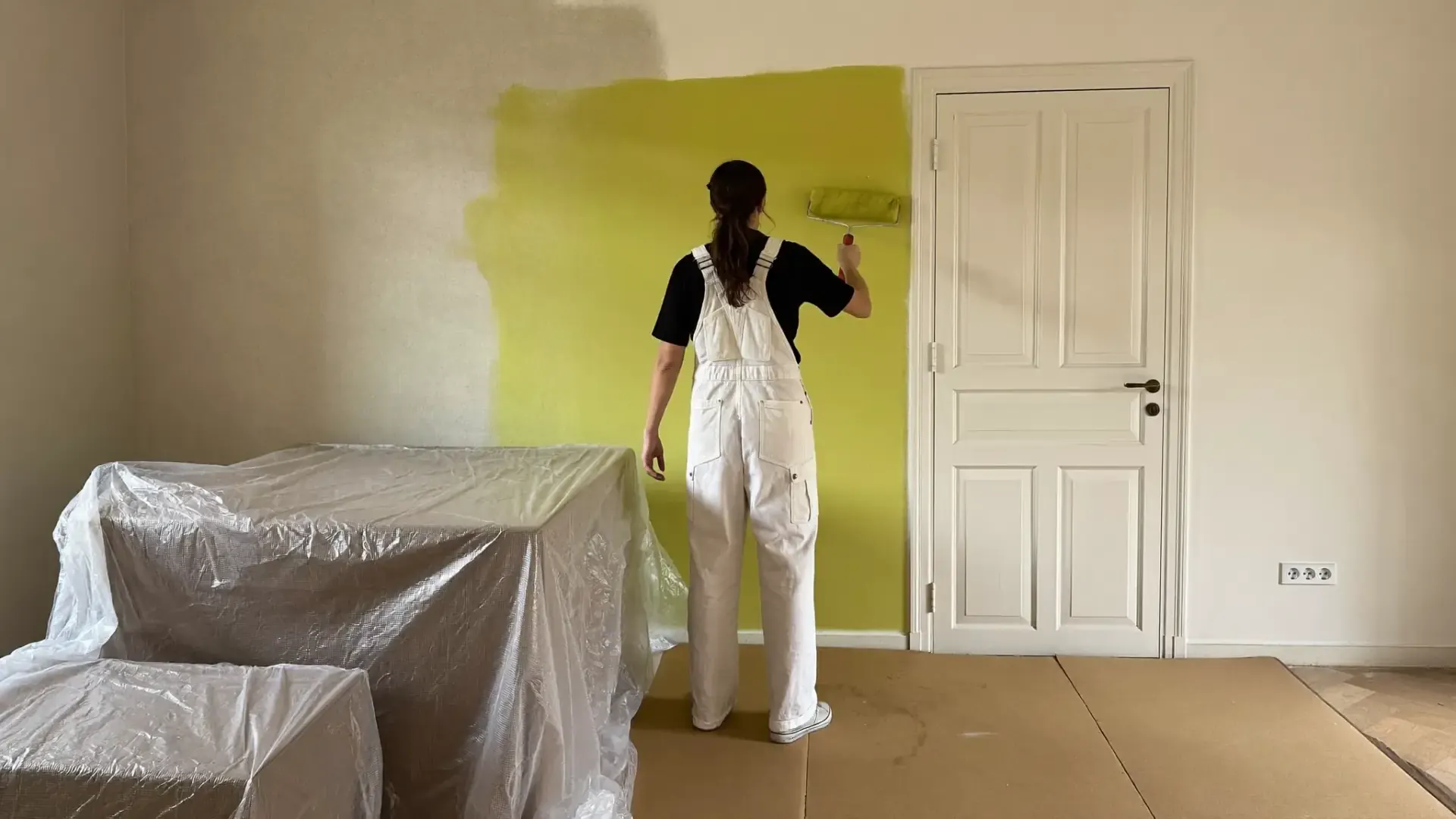 Pintor de espaldas pintando con rodillo, muebles protegidos con plástico
